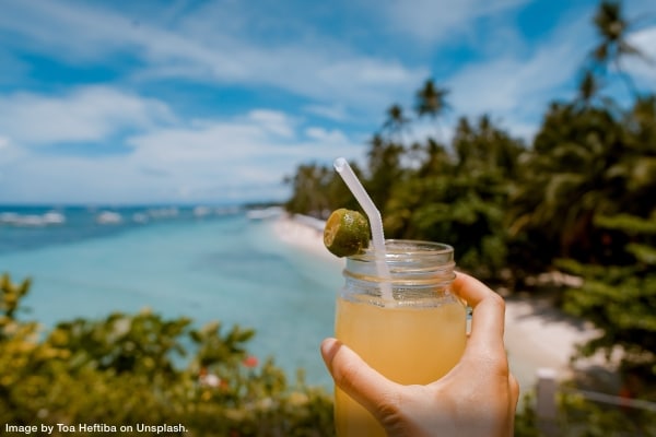 person's hand holding up a cocktail with a beach and ocean in the background representing vacation planning advice for entrepreneurs