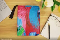 Zippered binder with multi-coloured cover on a desk beside a computer and notebook, pencil, eyeglasses and tulip in vase