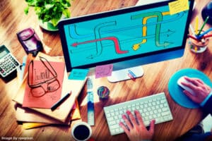 person in front of computer with one hand on keyboard other hand on mouse. desk piled with papers representing how to multitask successfully